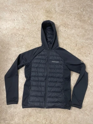 Svart Peak Performance pufferjacka - Svart pufferjacka från Peak Performance med huva och hel dragkedja. Jackan har quiltade partier framtill och baktill, samt släta ärmar. Snygg minimalistisk design med logga på bröstet och ärmen. Perfekt för kyliga dagar.