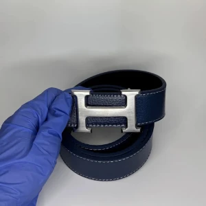 Marinblå H skärp i skinn - Snyggt marinblått bälte från H med klassisk H-spänne i silverfärgad metall. Bältet är tillverkat i exklusivt skinn med vit kontrastsöm längs kanterna. Perfekt accessoar för att lyfta din outfit med en lyxig touch.