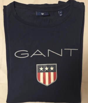 Marinblå långärmad GANT tröja - Snygg marinblå långärmad tröja från GANT med stort tryck på bröstet och sköld med stjärnor och rödvita ränder. Rund hals och mjukt bomullsmaterial gör den skön att bära. Perfekt för dig som gillar klassisk och stilren design.