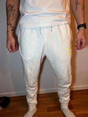 Vita Nike mjukisbyxor med broderi - Säljer ett par vita mjukisbyxor från Nike med broderade neongula swoosh-loggor och blå textdetaljer längs benet. Byxorna har snörning i midjan, ribbade muddar och sidofickor. Perfekt för chill eller träning.
