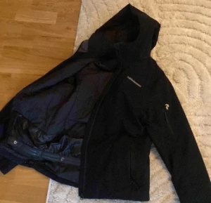 Svart vindjacka från Peak Performance - Svart vindjacka från Peak Performance med huva och flera praktiska fickor med dragkedja. Jackan har en stilren design och är tillverkad i slitstark polyester, perfekt för blåsiga dagar. Snygg logga på bröstet och ärmen, samt justerbar nederkant.