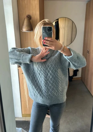 Grå stickad tröja med kabelmönster - Mysig grå stickad tröja med klassiskt kabelmönster framtill. Tröjan har en rund halsringning och långa ärmar. Passformen är loose.