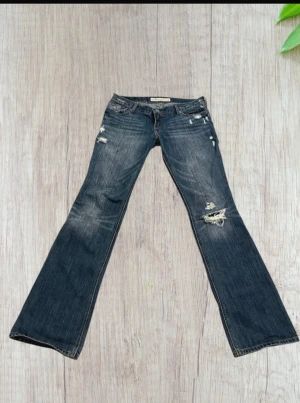 Blå bootcut jeans från Hollister - Säljer ett par blå jeans från Hollister med bootcut passform och slitna detaljer. Jeansen har låg midja, klassisk femficksdesign och är tillverkade i jeansmaterial med snygga slitningar och hål på benen för en cool look. Midjemått = 41,5 och innerbenslängden = 81