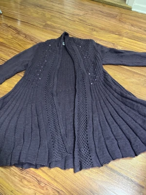 Mörklila stickad kofta från Isolde - Säljer en mörklila stickad kofta från Isolde i storlek L. Koftan har långärmade armar, öppet framstycke med breda virkade kanter och snygga paljettdetaljer på axlar och bröst. Plisserad nederdel ger extra schysst fall och cool look.