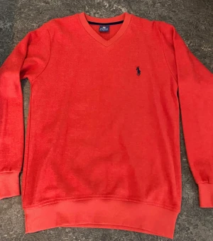 Röd v-ringad tröja från Polo Ralph Lauren - Snygg röd tröja från Polo Ralph Lauren med v-ringad hals och klassisk blå logga broderad på bröstet. Tröjan har långa ärmar och ribbade muddar vid hals, ärmslut och nederkant. Perfekt för dig som gillar stilrena och tidlösa plagg.