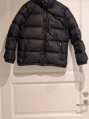 Svart pufferjacka Polo Ralph Lauren - Svart pufferjacka från Polo Ralph Lauren med röd logga på bröstet. Jackan har hög krage, tre dragkedjeförsedda fickor fram och elastiska muddar vid ärmslut. Materialet är syntet och jackan är quiltad för extra värme. Perfekt för kalla dagar.
