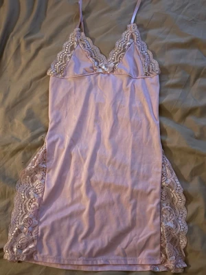 Ljusrosa nattlinne med omlott - Säljer ett ljusrosa nattlinne med tunna axelband och omlott-design. Linnet har spetsdetaljer längs sidorna och vid bysten, samt en liten rosett framtill. Materialet är mjukt och stretchigt, troligen polyester eller bomull, och ger en feminin och söt vibe.