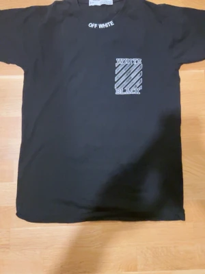 Svart Off-White t-shirt med tryck - Svart t-shirt från Off-White med vit text på kragen och coolt grafiskt tryck på bröstet och ryggen. Framsidan har ett randigt mönster med texten 'WHITE BLACK', medan ryggen har stora vita pilar och 'WHITE'-text. Klassisk passform och rund hals.