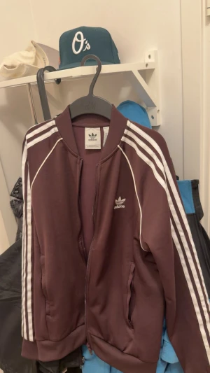  Adidas ziptröja med vita ränder - Säljer en Adidas ziptröja med klassiska vita ränder längs ärmarna och Adidas-logga på bröstet. Tröjan har dragkedja framtill, ribbade muddar och två fickor. Perfekt för en sportig och avslappnad stil.