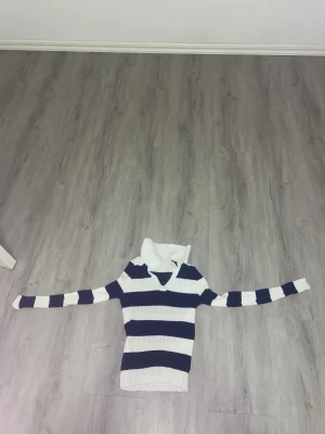 Randig polotröja i blått och vitt - Säljer en ribbstickad polotröja med breda marinblå och vita ränder. Tröjan har lång ärm, hög krage och en snygg v-ringning. Perfekt för dig som gillar klassisk stil med en twist. Storleken är L men passar även till M