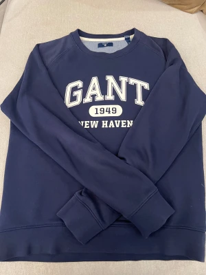 Mörkblå sweatshirt från GANT - Säljer en mörkblå sweatshirt från GANT med vit text 'GANT 1949 NEW HAVEN' på bröstet. Klassisk rund halsringning och långa ärmar. Tillverkad i mjuk bomull för skön känsla och stilren look.