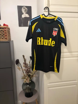 Adidas Como 1907 fotbollströja M - Snygg svart Adidas fotbollströja för Como 1907 med gula och blå detaljer, tryckt Rhude-logga och klubbmärke på bröstet. Slim fit-modell i lätt polyester som andas, perfekt för dig som gillar sportig stil och vill sticka ut på planen.