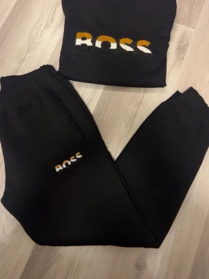 Hugo boss dress  - Svarta mjukisbyxor och tröja  från BOSS med broderad logga i vitt, orange och svart på benet. Byxorna har en avslappnad passform och är gjorda i mjukt material som är skönt mot huden. Perfekta för chill dagar och har en modern look.