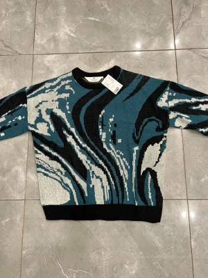 stickad tröja från H&M för pojke - Cool stickad tröja från H&M med abstrakt mönster i blått, svart och vitt. Tröjan har rund halsringning, ribbade muddar och en loose fit som ger en avslappnad vibe. Den är helt oanvänd med lappen på. 