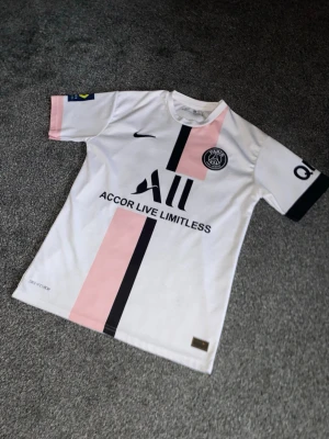PSG - Säljer en Paris Saint-Germain matchtröja från Nike i storlek S, med Messi 30 på ryggen. Tröjan är vit med rosa och svarta ränder, PSG-logga på bröstet och Dri-FIT material som andas. Snygg detalj med sponsortryck och svarta muddar på ärmarna.