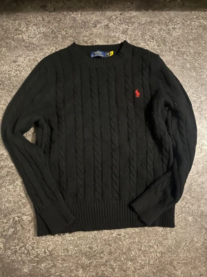 Svart kabelstickad tröja Polo Ralph Lauren - Svart kabelstickad tröja från Polo Ralph Lauren med klassisk röd broderad logga på bröstet. Tröjan har rund halsringning, ribbade muddar och är långärmad. Perfekt för dig som gillar stilren och tidlös design.