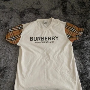 Vit Burberry t-shirt med rutiga ärmar - Säljer en snygg vit t-shirt från Burberry med klassiska rutiga kortärmar i beige, svart och rött. Stor Burberry-logga på bröstet. T-shirten har rund hals och är gjord i mjuk bomull. Perfekt för dig som gillar designer och vill sticka ut.