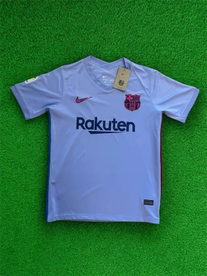 Barça Nike matchtröja lila - Säljer en lila FC Barcelona matchtröja från Nike med Rakuten-tryck på bröstet och klubbmärke. Tröjan har kort ärm, V-ringning och detaljer i rött och blått längs sidorna. Materialet är lätt och andas, perfekt för fotboll eller att bära som supporter.