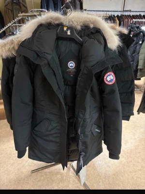 Svart dunjacka från Canada Goose - Säljer en svart dunjacka från Canada Goose med fet pälskantad huva och klassisk logga på ärmen. Jackan har dragkedja, knappar och flera fickor framtill. Perfekt för kalla vinterdagar och riktigt snygg streetstil.