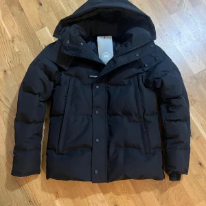 Canada Goose wyndham black label - Storlek M. Normal fit. Aldrig använd. Säljer en svart dunjacka från Canada Goose med huva och snyggt broderat märke på ärmen. Jackan har dragkedja och knappar framtill, samt två stora fickor. Perfekt för kalla vinterdagar och har en riktigt schysst, puffig look. 
