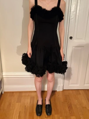 Svart volangklänning med spetsdetaljer - Vacker svart klänning med breda axelband och volangdetaljer vid halsringning och nederkant. Klänningen har en figurnära passform upptill och ett fluffigt, voluminöst kjolparti med spets. Dragkedja baktill för enkel påklädning. Säljes då den är något för stor på mig. 🖤