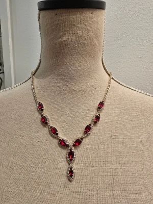 Röd glittrig statement-halsband - Säljer ett riktigt snyggt halsband med stora röda stenar och glittriga detaljer. Kedjan är guldfärgad och stenarna är formade som droppar, vilket ger en lyxig och elegant vibe. Perfekt för att lyfta vilken outfit som helst.