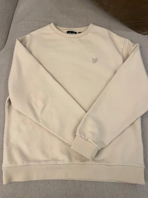 Beige sweatshirt från Lyle & Scott - Säljer en stilren beige sweatshirt från Lyle & Scott med diskret broderad logga på bröstet. Tröjan har rund halsringning, långa ärmar och ribbade muddar. Perfekt för dig som gillar enkel och clean stil.