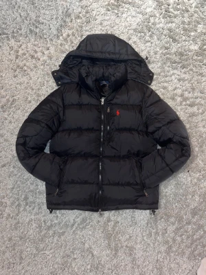 Svart dunjacka från Polo Ralph Lauren - Svart dunjacka från Polo Ralph Lauren med huva och röd logga på bröstet. Jackan har quiltad design, dragkedja framtill och två sidofickor med dragkedja. Perfekt för kalla dagar och har en clean, sportig look.