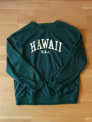 Grön sweatshirt med tryck - Mörkgrön sweatshirt med "HAWAII" tryckt på bröstet. Tröjan har rund halsringning, långa ärmar och ribbade muddar vid ärmslut och nederkant. Perfekt för en chill och avslappnad stil.