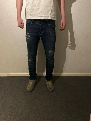 Jack & Jones Slim Glenn jeans blå - Säljer ett par mörkblå slim fit jeans från Jack & Jones, modell Slim/Glenn. Jeansen har slitna detaljer och snygga slitningar. Jeansen är knappt använda men har en slitning som öppnats vid bak fickan som man ser på sista bilden.
