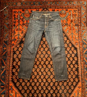 Mörkblå jeans från Nudie Jeans - Säljer ett par mörkblå jeans från Nudie Jeans med klassisk femficksdesign och kontrastsömmar. Jeansen har raka ben och snygga detaljer på bakfickorna med Nudie-loggan. Tillverkade i slitstarkt denimtyg av bomull.