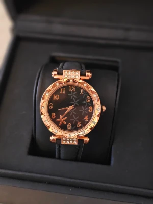 Svart och guld klocka med blommönster - Snygg klocka med svart urtavla och blommönster, omgiven av en guldig ram med glittrande stenar. Armbandet är svart på ovansidan och beige på undersidan, gjort i syntetmaterial. Siffrorna är i guld och ger en lyxig vibe. Perfekt accessoar för att lyfta din stil.