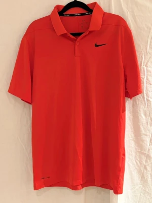 Röd Nike piké stl M - Säljer en röd Nike piké i  Dri-Fit material med klassisk krage och korta ärmar. Tröjan har svart Nike-logga på bröstet och är tillverkad i ett lätt, snabbtorkande syntetmaterial som andas. Perfekt för golfbanan eller sportiga dagar.