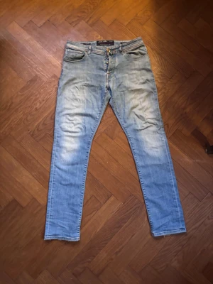 Jacob Cohën ljusblå jeans - Snygga ljusblå jeans från Jacob Cohën med klassisk femficksdesign och knappgylf. Jeansen har coola kontrastsömmar i orange och grönt, samt en unik grön-randig patch med J-logga bak. Perfekt passform och italiensk känsla.
