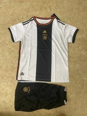 Tysklands fotbollsset Adidas svart/vit - Säljer ett officiellt Tyskland fotbollsset från Adidas med tröja och shorts. Tröjan är vit med svart mittfält, guldiga detaljer och Tysklands emblem med fyra stjärnor. Shortsen är svarta med samma emblem. Setet har ränder i tysklands färger längs sidorna och är gjort i lätt polyester.