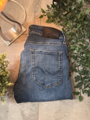 Blå jeans från Jack & Jones - Snygga blå jeans från Jack & Jones. Jeansen har ljusa slitningar, kontrastsömmar och läderpatch med logga bak i midjan. Materialet är mjukt denim med stretch för extra komfort. Skriv för funderingar!