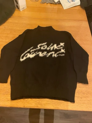 Svart stickad tröja med vit text saint Laurent  - Cool svart stickad tröja med vit, fluffig text framtill i grafisk stil. Tröjan har lång ärm, ribbade muddar och en något högre hals. Perfekt för dig som gillar statement-plagg och vill sticka ut lite extra.