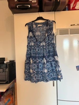 Blå mönstrad kortklänning Odd Molly - Supersnygg blå och vit kortklänning med paisley-inspirerat mönster och v-ringad hals. Klänningen är ärmlös och har tunna knytband vid halsen. Tillverkad i tunn och skön bomull, perfekt för varma dagar. Passformen är normal och tyget har en lätt, luftig känsla. Från märket Odd Molly. Storlek 2 som enligt deras storleksguide motsvarar en M. Mycket fint skick, jag hittar inga synliga anmärkningar eller defekter. 