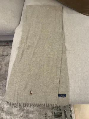 Beige kashmirhalsduk från Polo Ralph Lauren - Snygg beige halsduk från Polo Ralph Lauren i mjuk kashmir. Klassisk design med fransar i nederkant och broderad logga med polospelare. Perfekt för dig som gillar stilrena och lyxiga detaljer. Diskret Polo-märke i blått och gult på sidan.