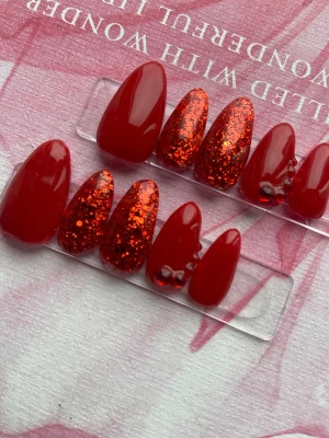 Röda glitter press-on naglar - Snygga press-on naglar i mandelform med intensivt röd färg och glittriga accenter. Några naglar har röda pärldekorationer för extra detalj. Perfekt för dig som vill ha en bold look med både glans och glitter. Materialet är gele och färgen är djup röd med -rött glitter.