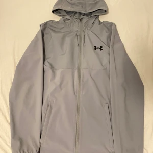 Grå vindjacka från Under Armour - Snygg grå vindjacka från Under Armour med huva och hel dragkedja framtill. Jackan har svarta loggor på bröstet och ryggen, två sidofickor och justerbar snörning vid huvan. Perfekt för dig som gillar sportig och clean stil.