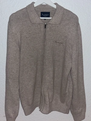 Beige stickad kofta från Bondelid - Säljer en beige stickad kofta från Bondelid med klassisk krage och hel dragkedja framtill. Diskret broderad logga på bröstet och ribbade muddar vid ärmslut och nederkant. Perfekt för lager-på-lager under kyliga dagar.