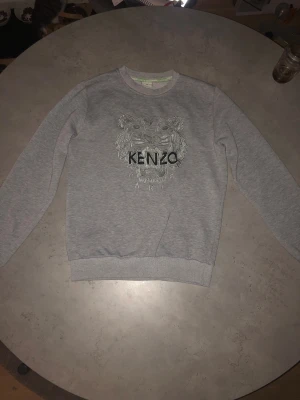 Grå sweatshirt från Kenzo med tiger - Grå sweatshirt från Kenzo i storlek M med ikonisk broderad tiger och Kenzo Paris-logga på bröstet. Tröjan har rund hals, ribbade muddar och är långärmad. Perfekt för dig som gillar streetstyle och vill sticka ut med ett snyggt statement plagg.(pris kan diskuteras)