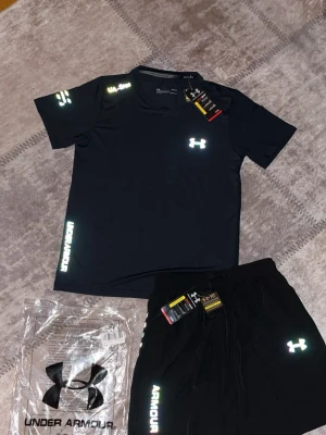 Under Armour reflactive set size xL  - Bilderna visar ett svart träningsset från Under Armour med t-shirt och shorts, båda med reflekterande loggor och tryck. Inga skor finns med på bilderna.