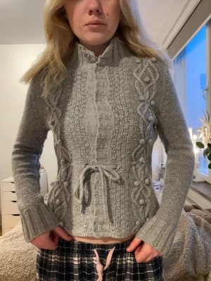 Grå stickad kofta med snörning - Säljer en grå stickad kofta med snygga flätade detaljer och bollar längs sidorna. Koftan har hög krage med knappar och ett knytband i midjan för extra stil. Långärmad och ribbade muddar vid ärmslut. Perfekt för dig som gillar mysiga och trendiga plagg.