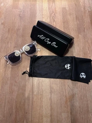 All cap one solglasögon  - Bilderna visar ett par transparenta solglasögon med svart fodral och putsduk från All Cap One, men ingen väst finns med.