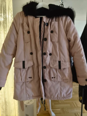 Beige parkajacka med svart fuskpäls - Snygg beige parkajacka med svart fuskpäls på huvan. Jackan har flera stora fickor med knappar, dragkedja och snörning i midjan för extra passform. Perfekt för kalla dagar med sin långa modell och fodrade insida. Stilsäker look med detaljer i svart.