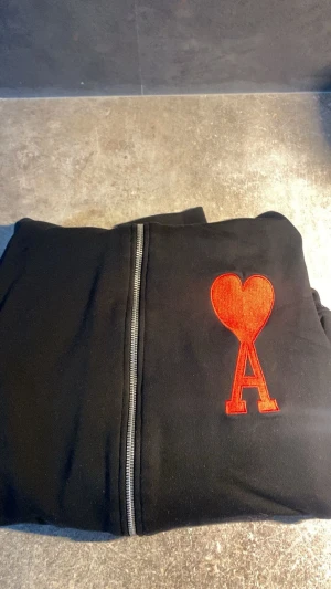 AMI PARIS - Svart jacka från Ami Paris i 100% bomull med dragkedja framtill. På bröstet sitter ett stort rött broderat hjärta med bokstaven A. Jackan har fickor på sidorna och en clean, stilren look som passar perfekt till streetwear.Storlek S 