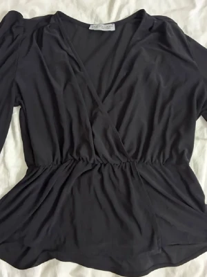 Svart omlottopp från Paul Mark - Svart omlottopp från Paul Mark i storlek XXL. Toppen har lång ärm, v-ringad omlottfront och ett insytt resårband i midjan som ger en snygg siluett. Materialet är mjukt och lätt, perfekt för dig som gillar stilrena och bekväma plagg.