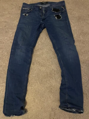Blå slitna jeans från Replay - Säljer ett par blå jeans från Replay med slitna detaljer och trasiga partier framtill. Jeansen har klassisk femficksdesign, raka ben och normal passform. Materialet är jeans i bomull och de har en snygg tvättad look.